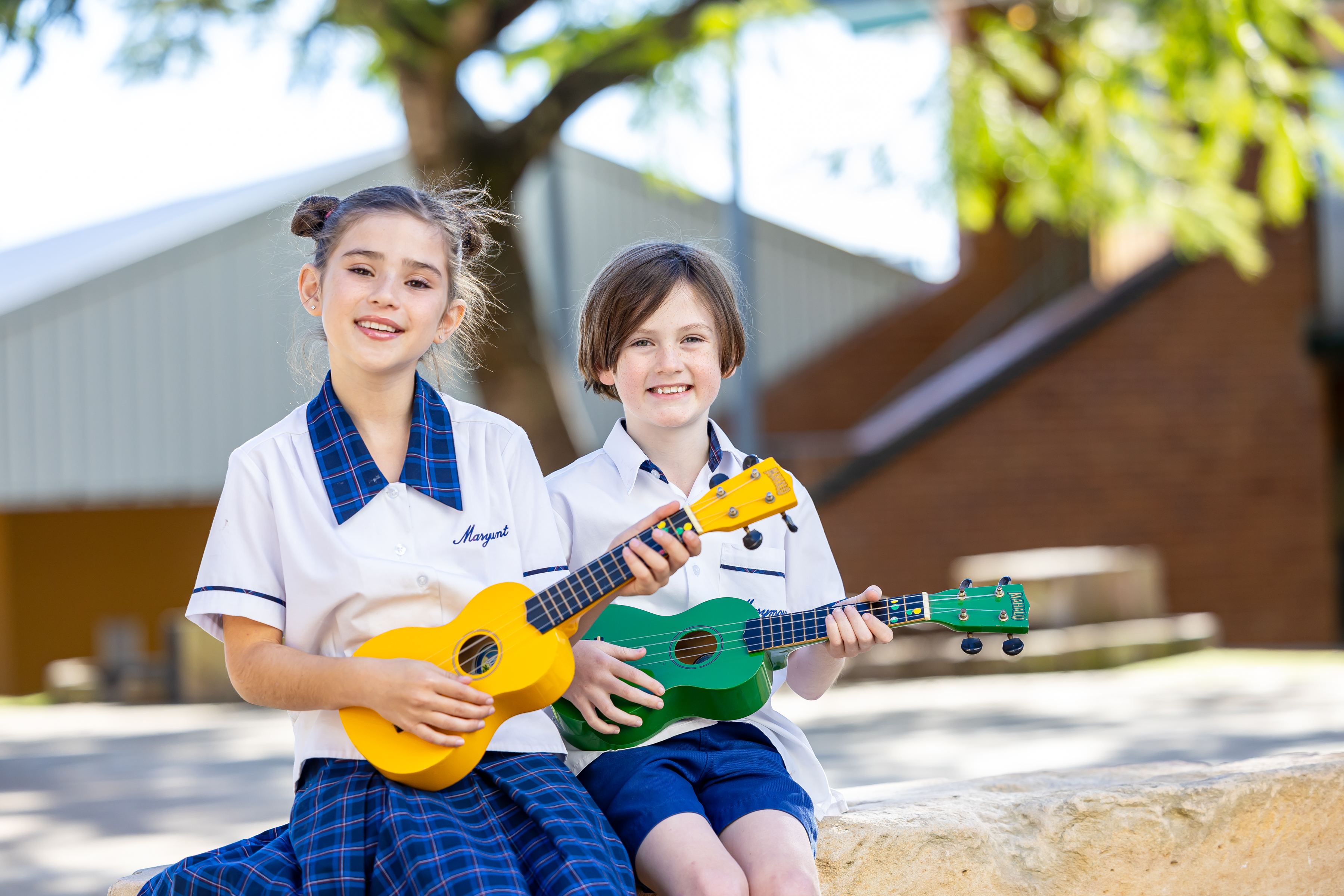 Two students in school uniforms sitting outdoors holding colourful ukuleles on a stone wall, with trees and school buildings in the background.