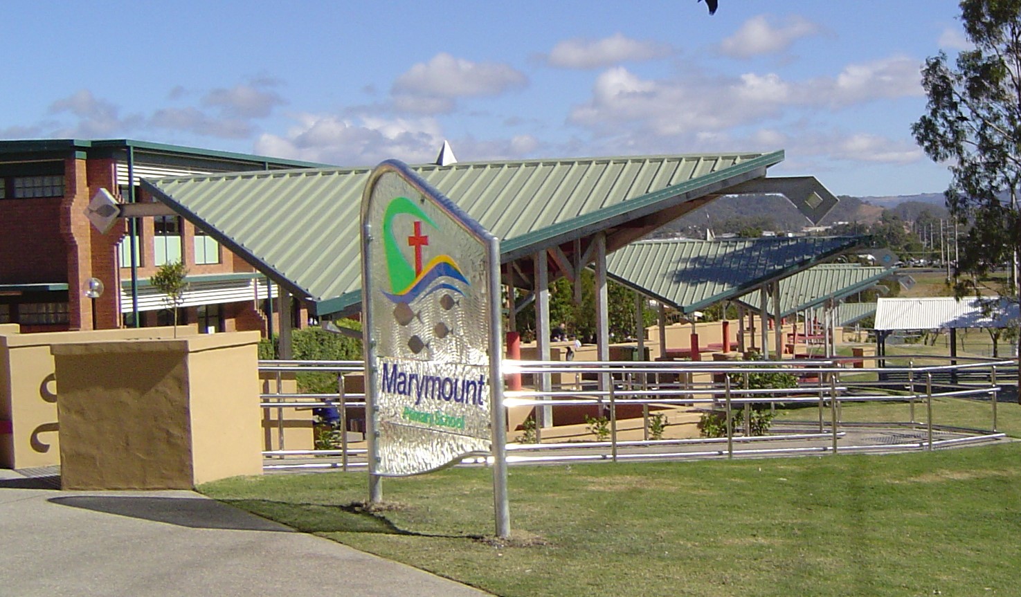 Sign reading “Marymount Primary School” in front of a school campus with covered walkways, railings, and buildings in the background.
