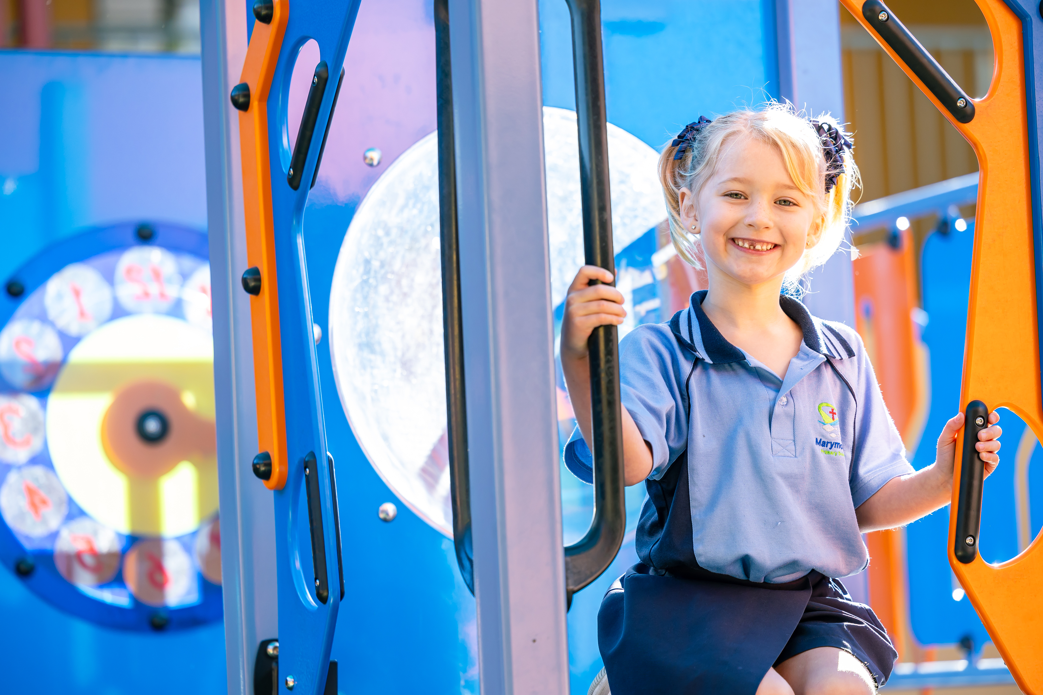 Student in a school uniform holding onto playground bars beside colourful play equipment outdoors.