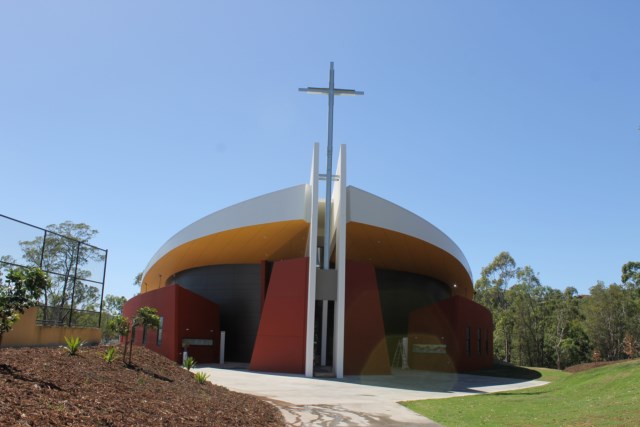 Modern circular chapel building with a large cross above the entrance, surrounded by landscaped grounds and trees.