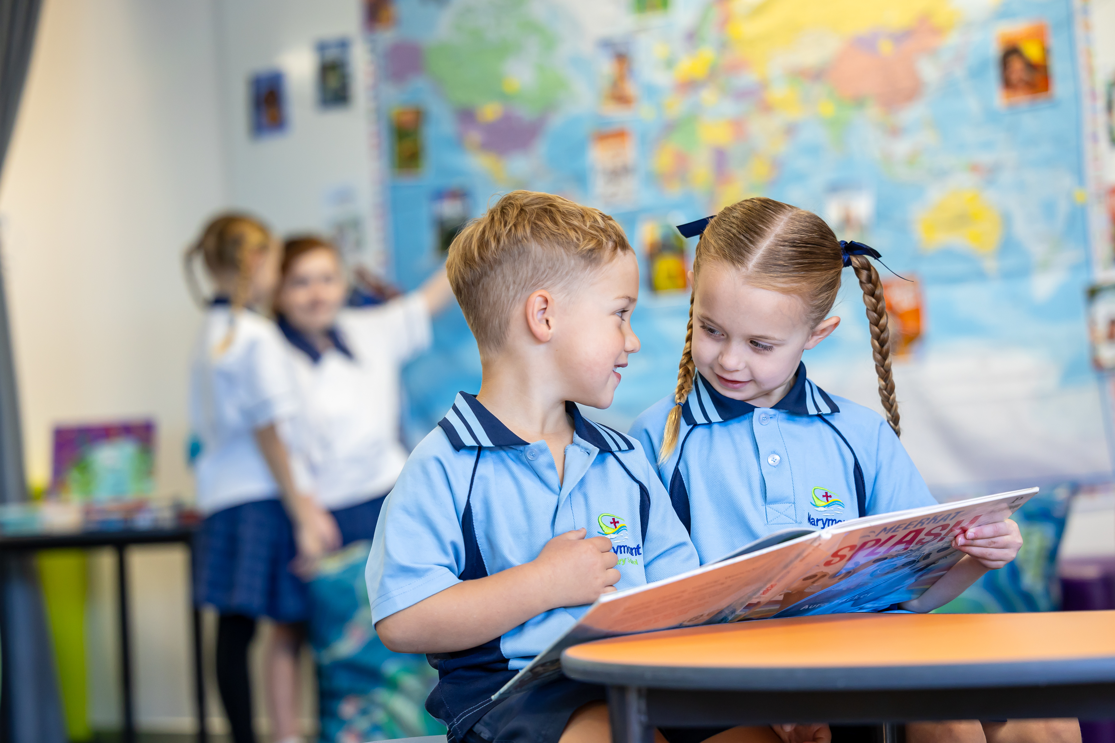 Two students in school uniforms reading a book together at a classroom table, with a world map and other students in the background.