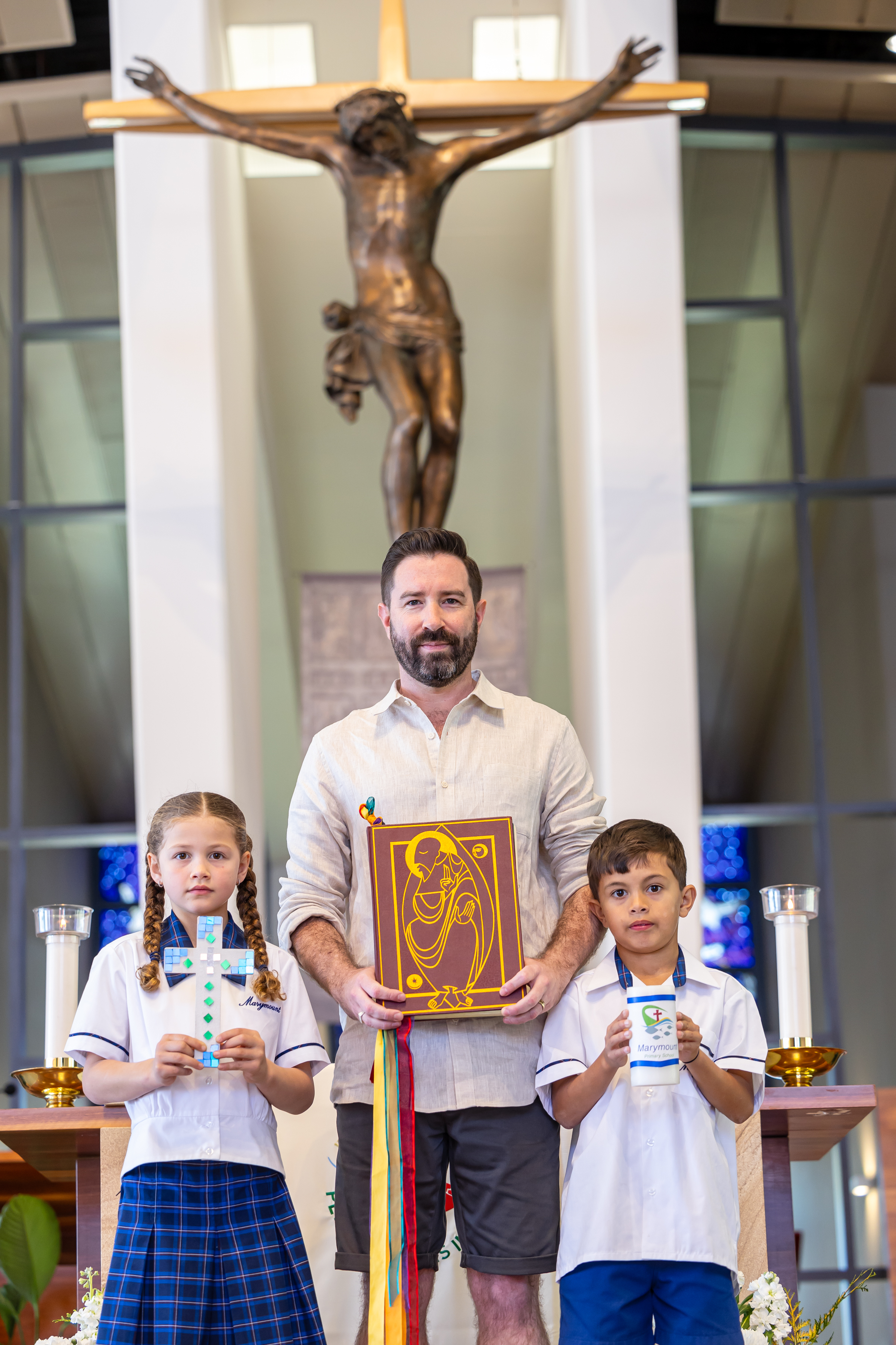 Two students and an adult standing at a church altar holding a cross, a book, and a candle, with a crucifix in the background.