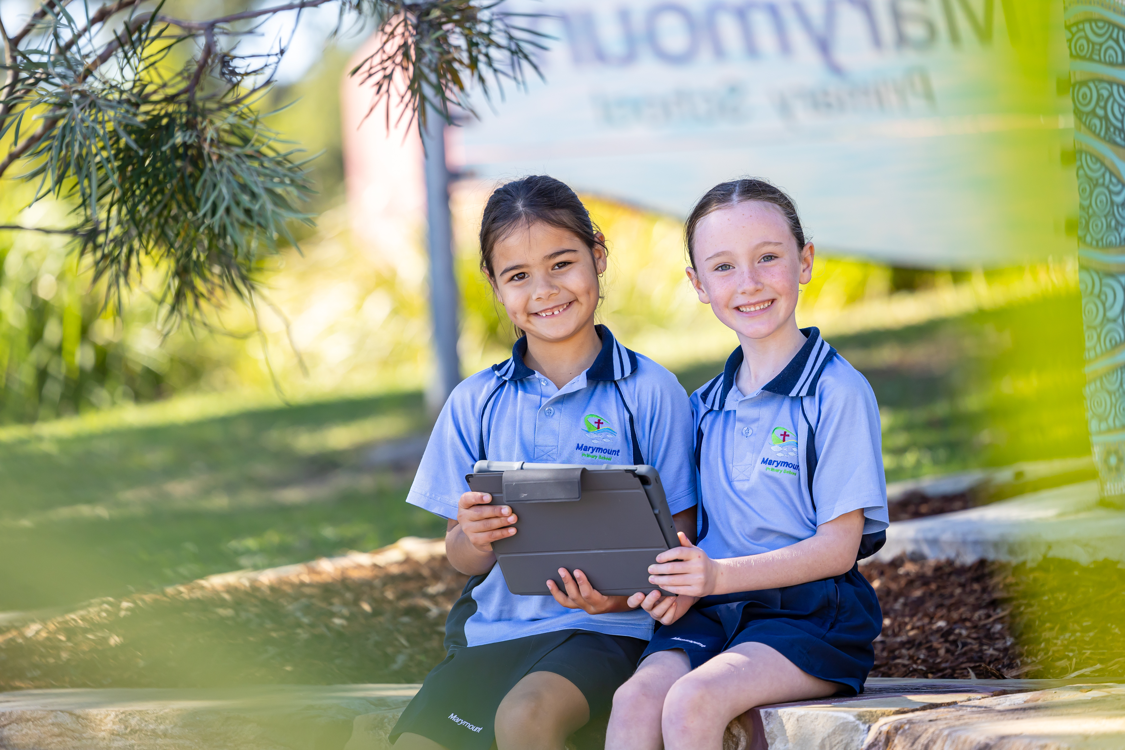 Two students in school uniforms sitting outdoors holding a tablet together on a stone wall, with trees and school signage in the background.