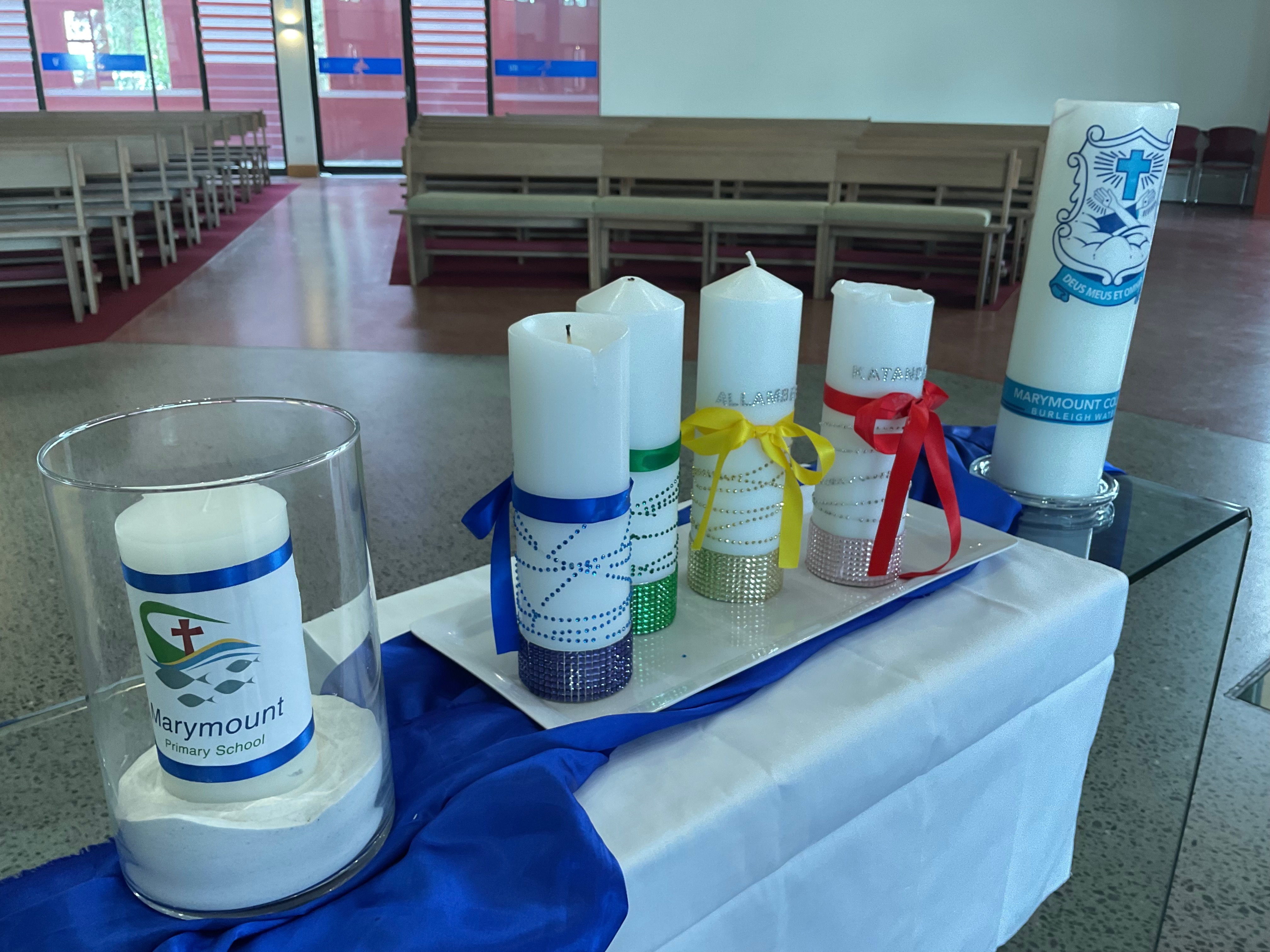 Display of decorated candles on a table inside a church or hall, including a Marymount Primary School candle and others wrapped with coloured ribbons.