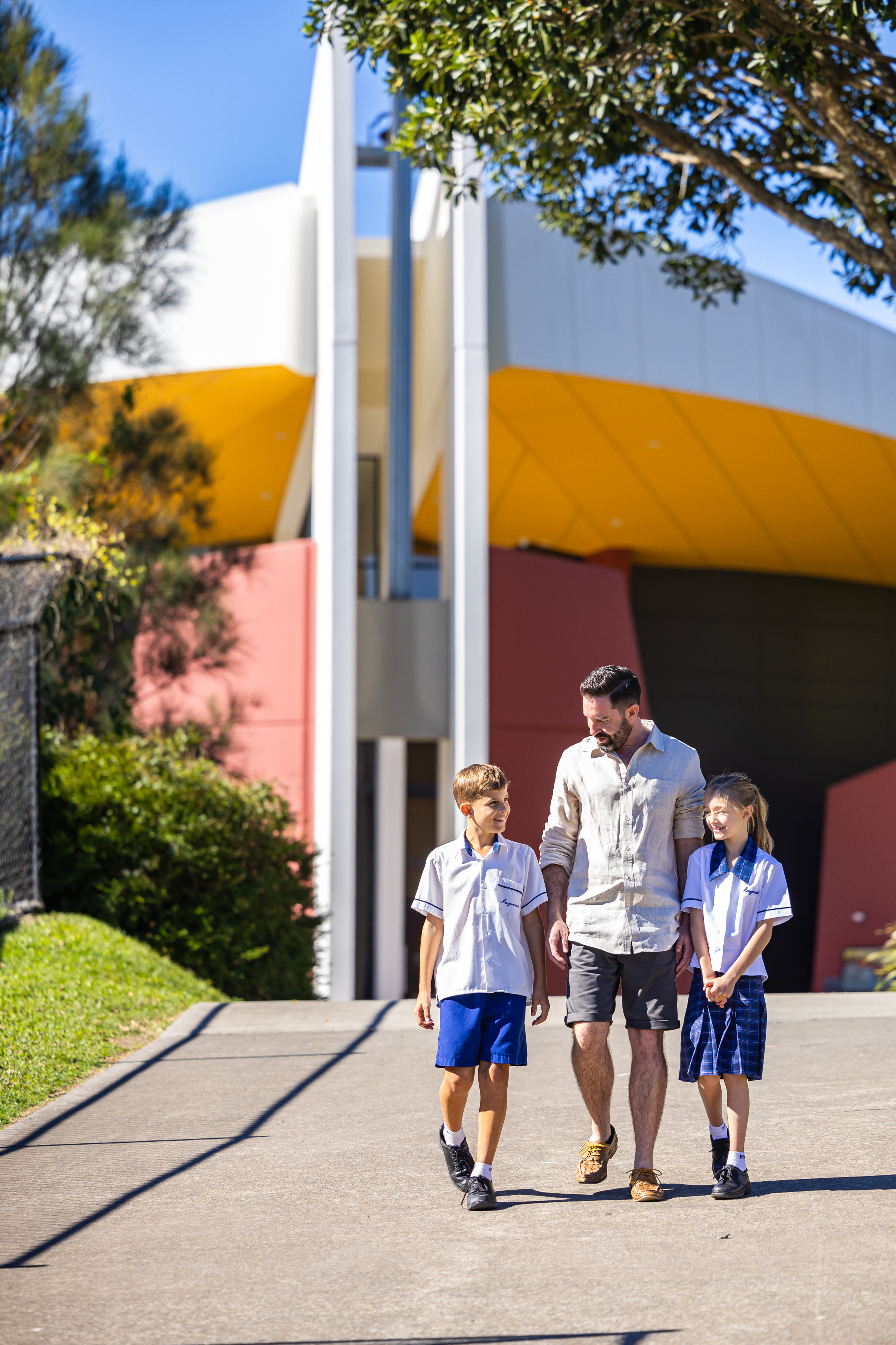 Adult walking with two students in school uniforms along a paved path in front of a modern school building.