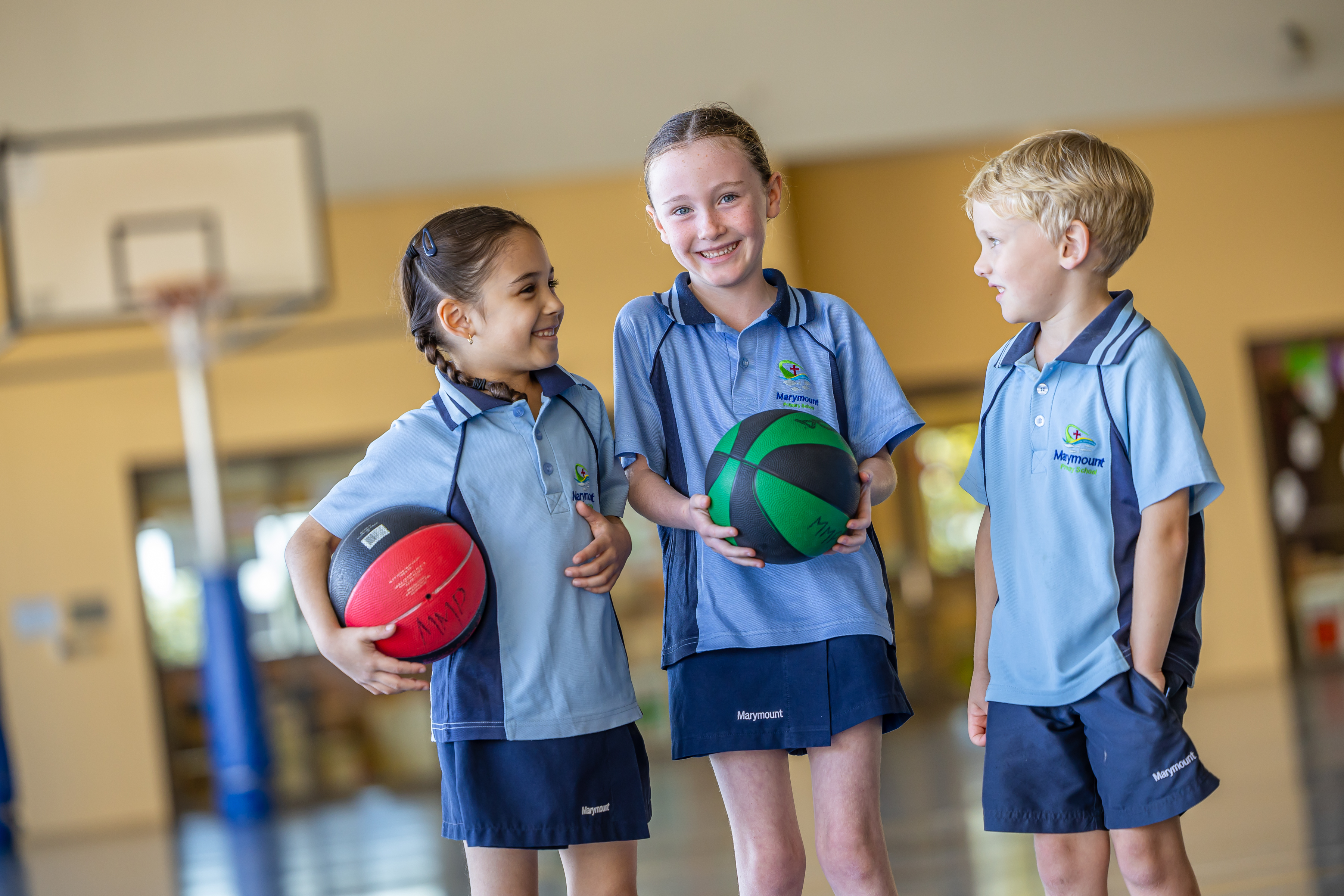 Three students in school sports uniforms standing indoors holding basketballs, with a basketball hoop visible in the background.