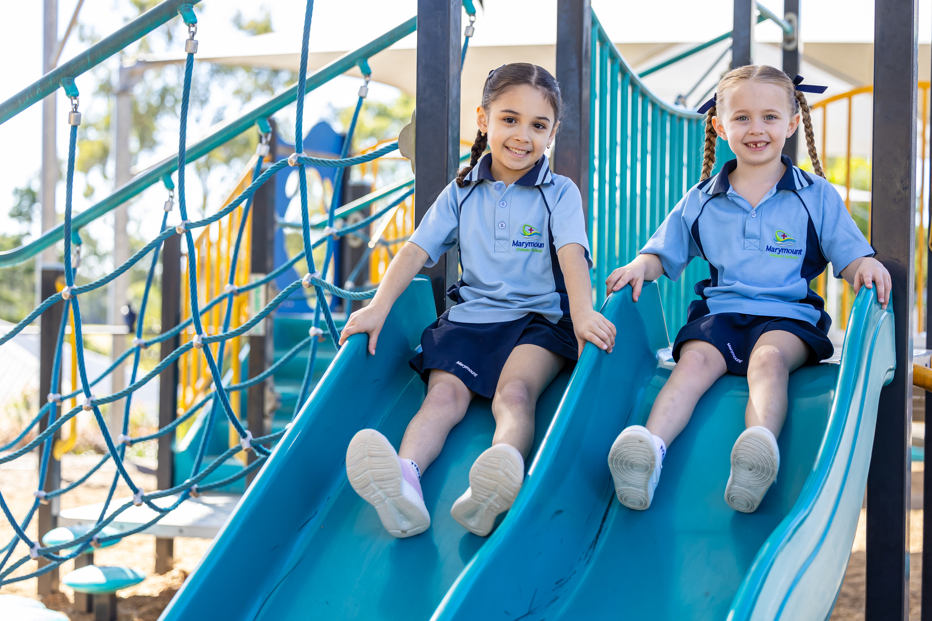 Two students in school uniforms sitting side by side on a playground slide, with climbing ropes and play equipment around them outdoors.